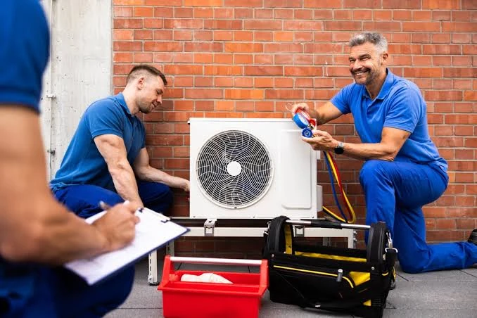 HVAC technicians inspecting a compact outdoor air conditioning system beside a brick wall